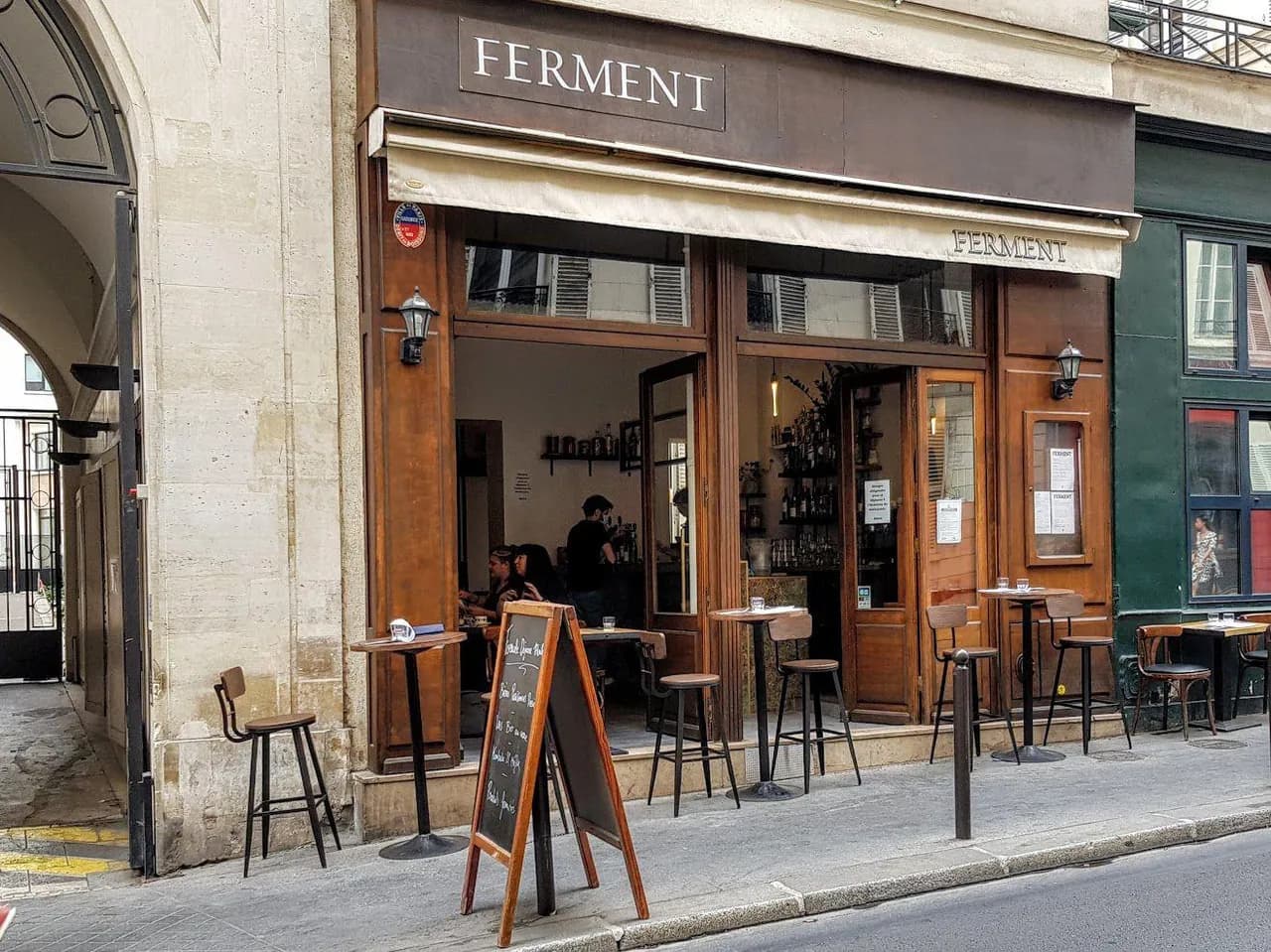 石造りのカフェの濃い色の木製看板。左は FERMENT、右は同じクリーム色のセリフ体で Delicious Treats に変わっています (元の写真)